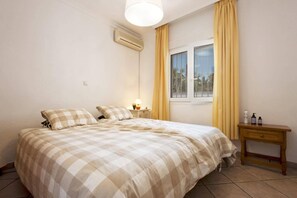 Ein Schlafzimmer mit Bett, Nachttisch, Fenster mit Vorhängen, Klimaanlage und einem kleinen Holztisch mit Pflegeprodukten.