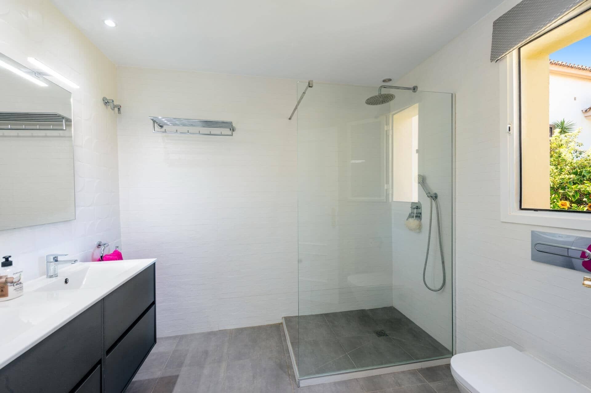 Un baño moderno con ducha envuelta en vidrio, un tocador blanco con alacenas oscuras y un espejo grande.