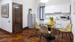 A compact kitchen with white cabinets, a refrigerator, and a microwave. There is a dining table with chairs and a vase with flowers.
