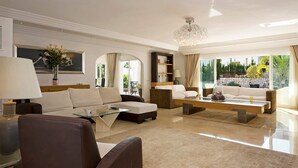 A spacious living room with a modern chandelier, a sofa set, a glass coffee table, and a view of a landscaped garden through large windows.