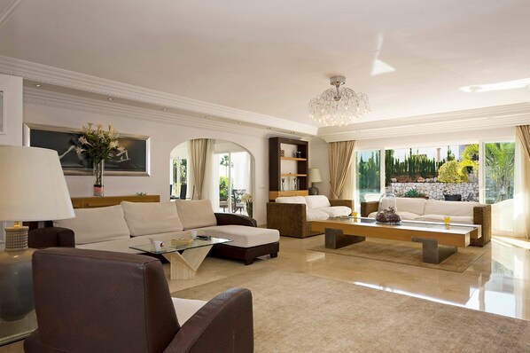 A spacious living room with a modern chandelier, a sofa set, a glass coffee table, and a view of a landscaped garden through large windows.