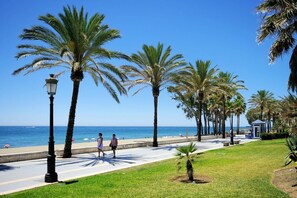 A coastal promenade with palm trees, a beach, and a small pavilion. - Villa Ramos by Villa Plus (Atalaya Isdabe)