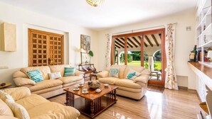 A spacious living room with a wooden coffee table, beige sofas, and a large window with a view of a garden. - Villa Ramos by Villa Plus (Atalaya Isdabe)