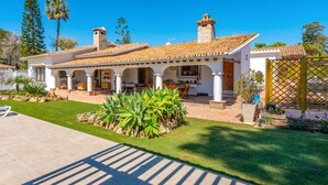 A two-story house with a tiled roof, a patio with a table and chairs, and a garden with large green plants. - Villa Ramos by Villa Plus (Atalaya Isdabe)
