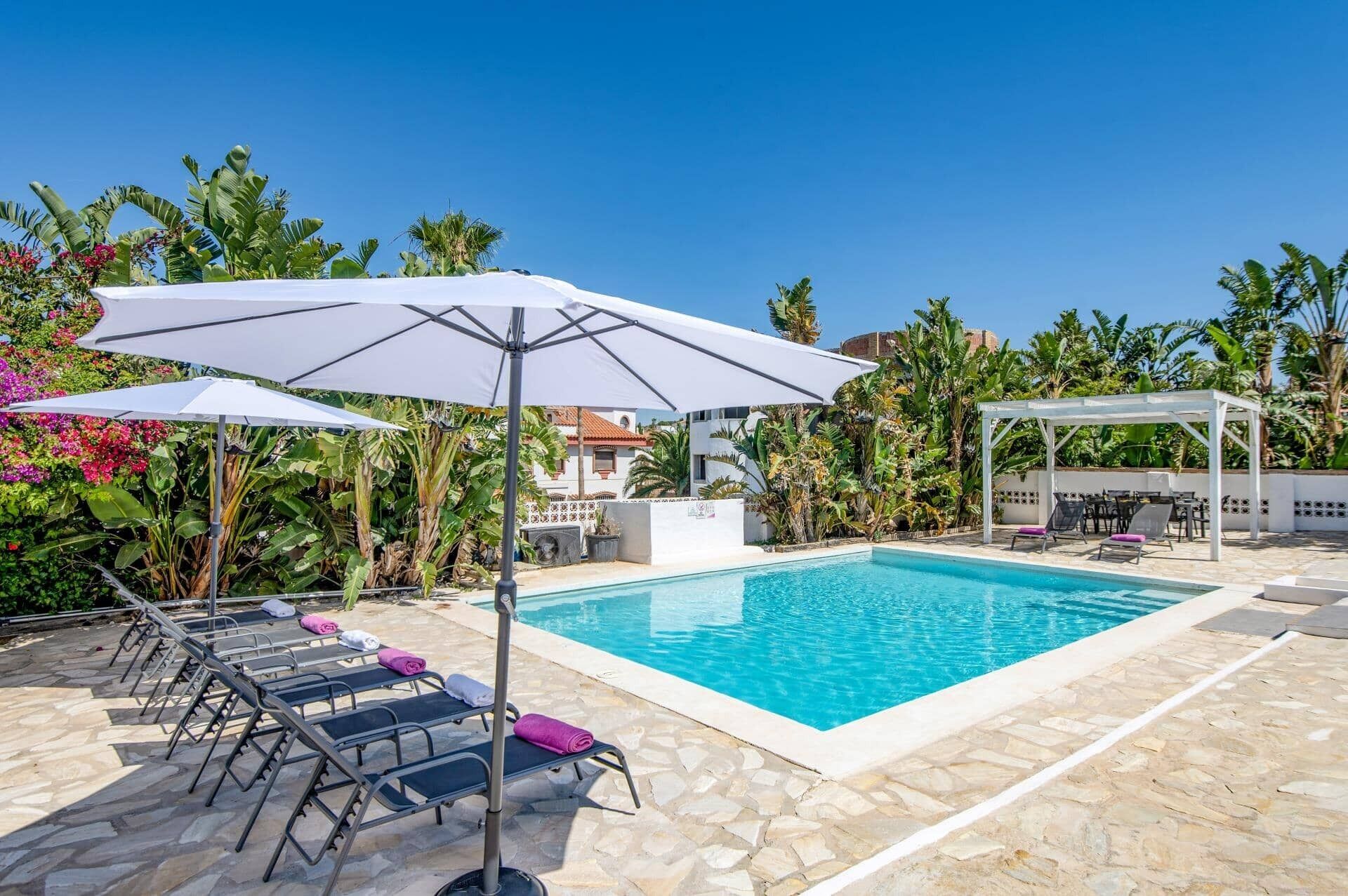 En swimmingpool med liggestole og parasoller, omgivet af tropiske planter og en hvid pergola.