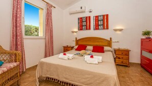 A bedroom with a wooden bed, red pillows, a wooden nightstand, a wicker chair, and a window with red curtains. - Villa Neptune by Villa Plus (Cap d'Artrutx)