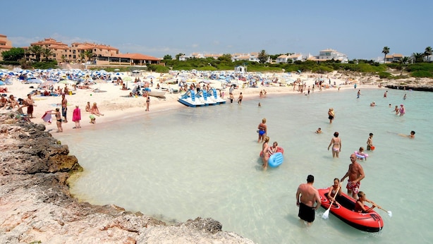 Ein überfüllter Strand mit badenden und sonnenden Menschen, eine felsige Küstenlinie und ein klarer, blauer Himmel.