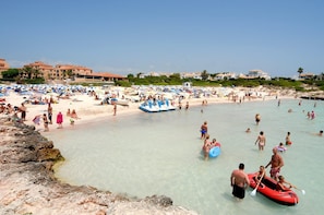 Una playa concurrida, gente nadando y tomando el sol, una costa rocosa y un edificio a lo lejos.