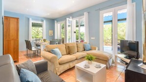 A spacious living room with a beige sofa, a coffee table, and a TV. There is a dining area with a table and chairs, and large windows offering a view of greenery. - Villa Serafina by Villa Plus (Son Parc)