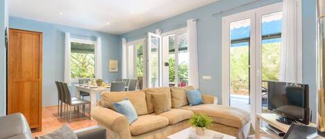 A spacious living room with a beige sofa, a coffee table, and a TV. There is a dining area with a table and chairs, and large windows offering a view of greenery.