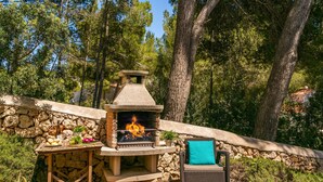 An outdoor stone fireplace with a fire, surrounded by a stone wall and a wicker chair with a cushion. - Villa Serafina by Villa Plus (Son Parc)