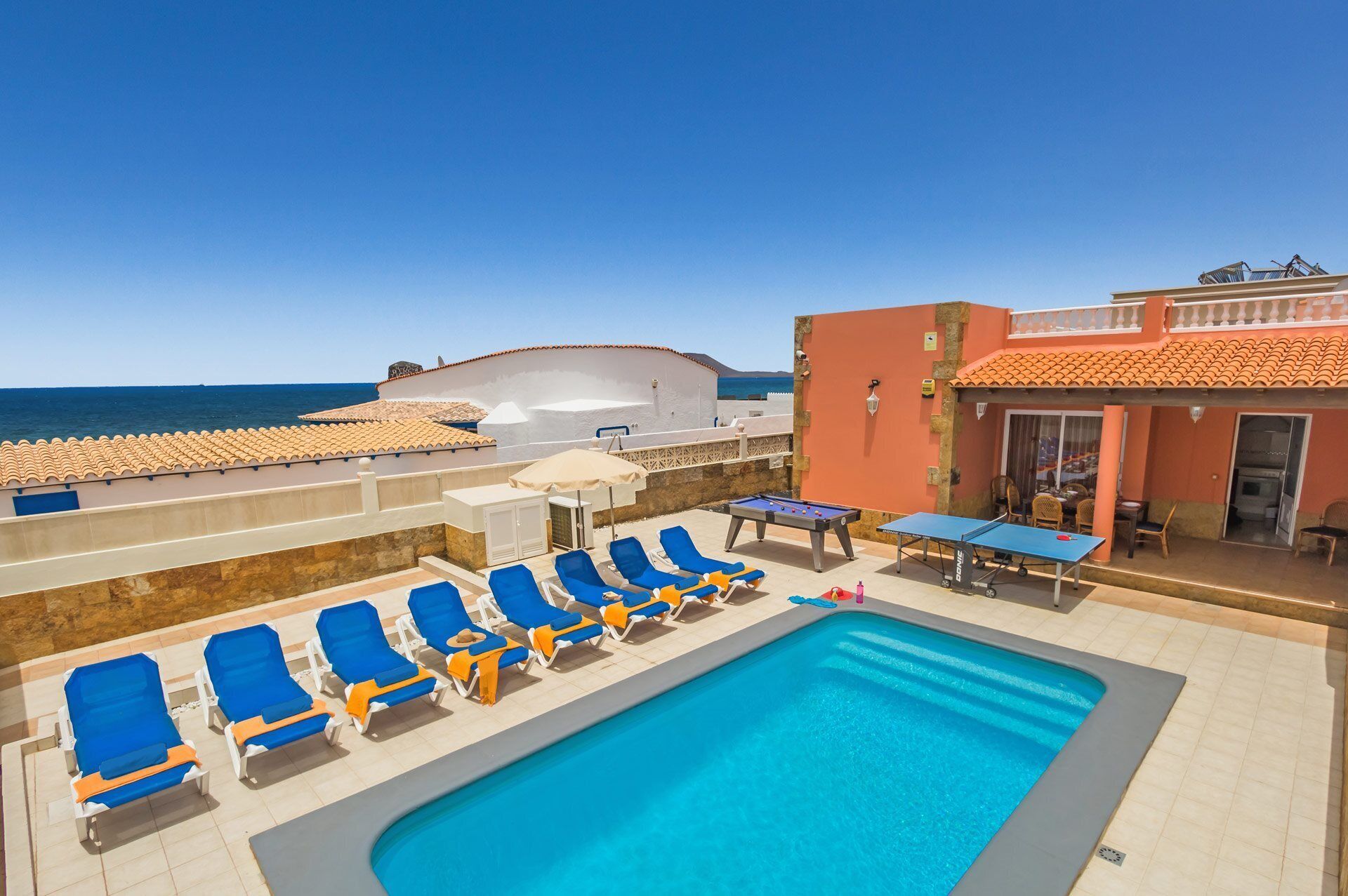 A pool area with blue lounge chairs and a table, a building with a red facade, and a view of the sea.