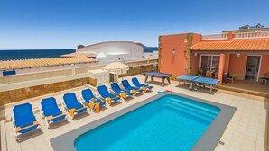 Ein Poolbereich mit blauen Liegestühlen und einem Tisch, ein Gebäude mit roter Fassade und Blick auf das Meer.
