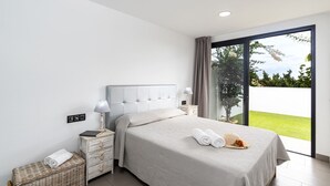 A modern bedroom with a large bed, a nightstand, a lamp, a small table with a book, and a wicker basket. - Villa Lola by Villa Plus (Corralejo)