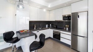 A modern kitchen with white cabinets, a stainless steel refrigerator, a microwave, and a built-in oven. There are two black bar stools and a black countertop. - Villa Lola by Villa Plus (Corralejo)