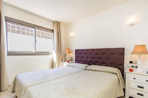Un dormitorio ordenado con una cama grande, una mesita de noche con lámpara y una ventana con un panel decorativo.