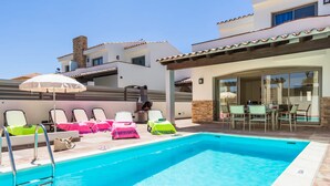 A swimming pool with lounge chairs and umbrellas, a patio area with tables and chairs, and a building with a tiled roof. - Villa Pino by Villa Plus (Corralejo)