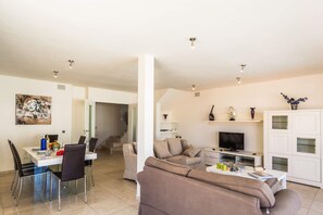 A modern living room with a sofa, TV, and a dining area with chairs and a table. - Villa Pino by Villa Plus (Corralejo)