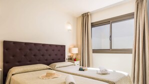 A hotel room with two beds, a large window with curtains, a bedside table with a lamp, and a wall-mounted phone. - Villa Pino by Villa Plus (Corralejo)