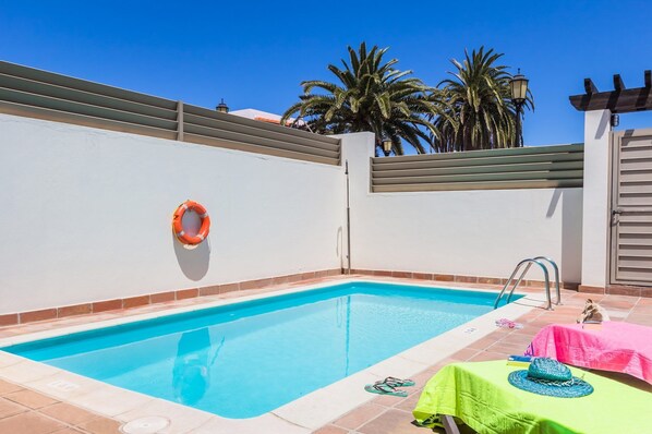 Une piscine rectangulaire bordée de carreaux, une bouée de sauvetage fixée au mur, une échelle en métal et une table rose avec une serviette verte et un chapeau.