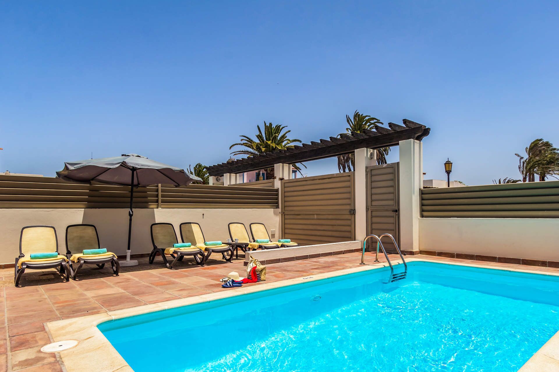 Una piscina con sillas de descanso y sombrillas, rodeada por un muro alto y palmeras.