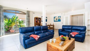 A living room with two blue sofas, a wooden coffee table with fruits and drinks, and a view of a balcony with plants. - Villa Jenny by Villa Plus (Corralejo)