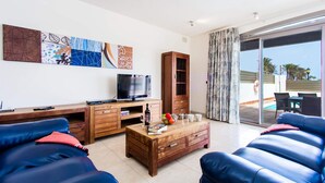 A modern living room with a TV, wooden furniture, and a view of a pool. - Villa Jenny by Villa Plus (Corralejo)