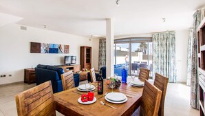 A dining area with a wooden table set for four, a television, and a sofa in the background. - Villa Jenny by Villa Plus (Corralejo)