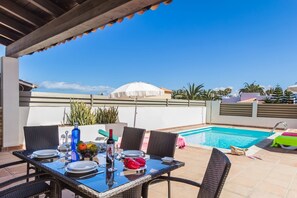 A patio with a pool, dining table set with plates, glasses, and a bottle, surrounded by wicker chairs.
