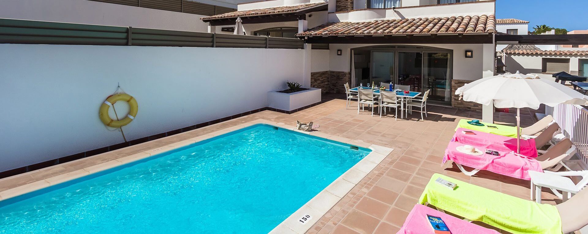 A swimming pool with lounge chairs and umbrellas, a patio area with a table and chairs, and a lifebuoy on the wall.