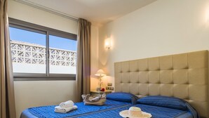 A hotel room with a bed, a nightstand, a lamp, a window with curtains, and a view of a building outside. - Villa Sirena by Villa Plus (Corralejo)