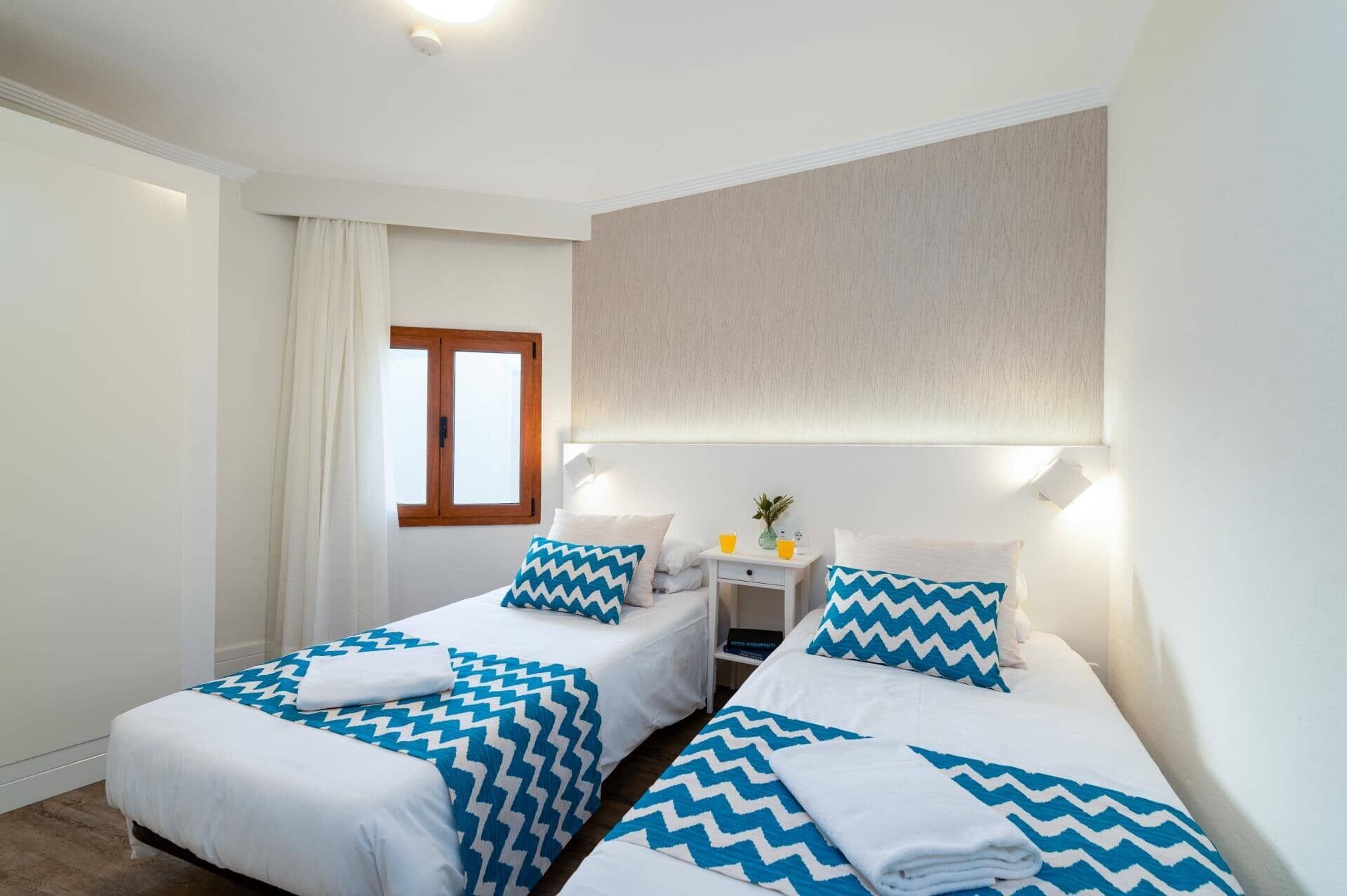 A room with two beds, each with blue and white zigzag patterned bedding, a small bedside table with a lamp, and a window with curtains.