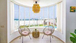 Een hoekzaal met een groot raam dat uitkijkt op een zwembad en strand. Twee rieten stoelen met kussens en een kleine ronde tafel met drankjes.