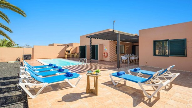 A pool area with lounge chairs and a small table, a covered patio, and a building with a flat roof. - Villa Palm View by Villa Plus (Corralejo)