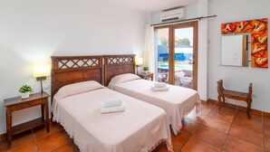 Une chambre d’hôtel avec deux lits, une tête de lit en bois, une table de chevet avec une lampe, une petite plante et une vue sur une piscine, visible à travers une porte ouverte.