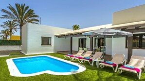 Ein moderner Poolbereich im Freien mit Liegestühlen und Sonnenschirmen, umgeben von einer gepflegten Rasenfläche und Palmen.