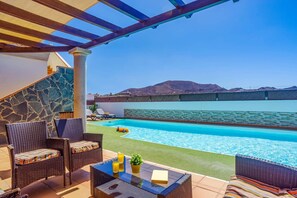 A poolside area with wicker furniture, a glass table, and a view of a large pool and mountains in the background.