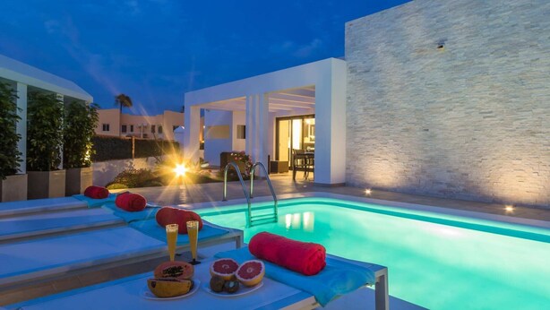Ein moderner Poolbereich im Freien mit Liegestühlen, einem Tisch am Pool mit Obst und Getränken und einem gut beleuchteten Innenraum, der durch die offene Tür sichtbar ist.