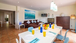 A modern living room with a large TV, a dining table set for two, and a kitchen area visible in the background.