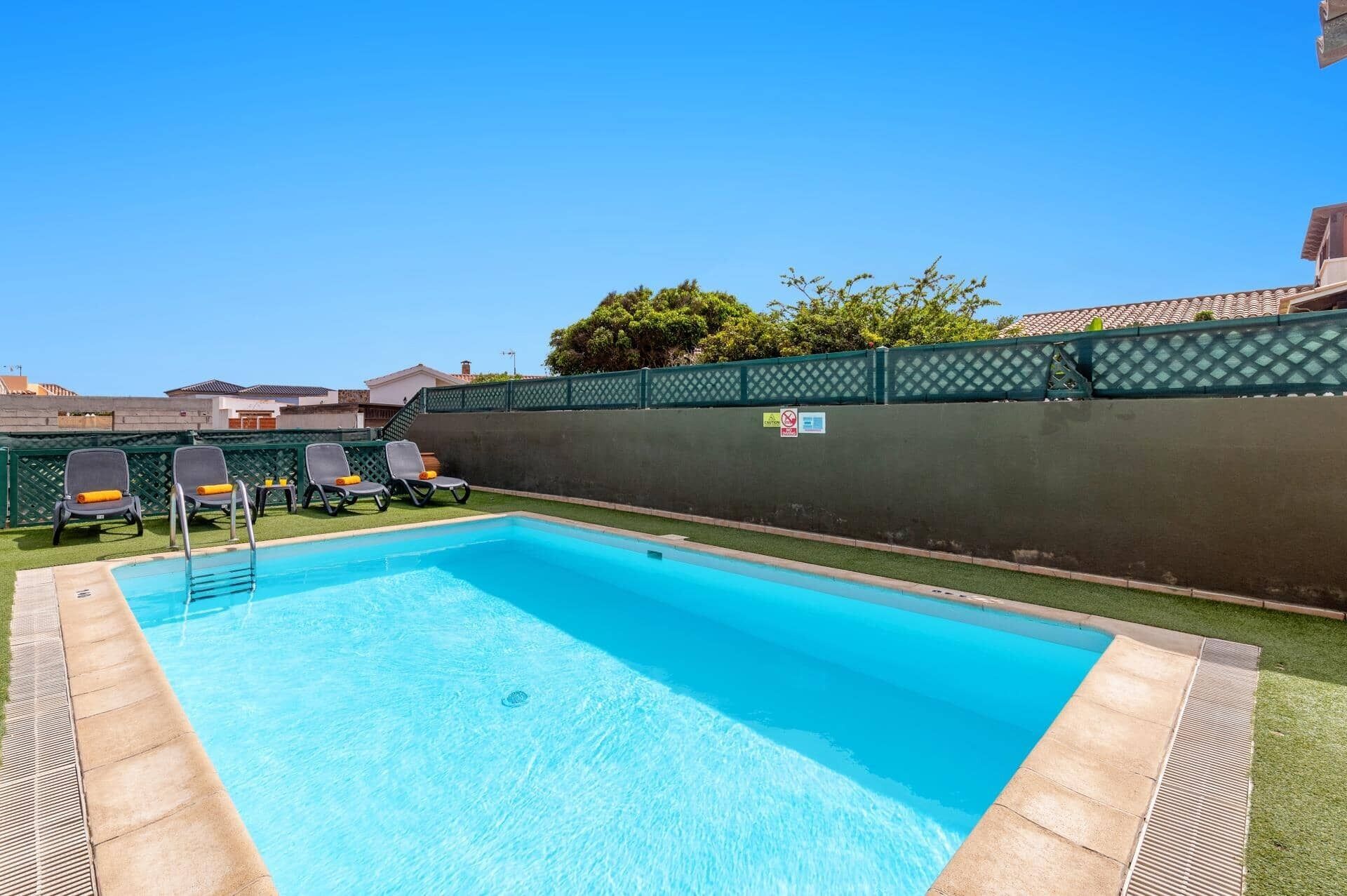 A rectangular swimming pool with a clear blue water surface, surrounded by a green fence and a grassy area. There are two lounge chairs with orange cushions and a pool ladder.