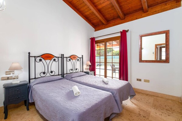 A double room with two beds, a wooden ceiling, a window with red curtains, a mirror, and a small bedside table with a lamp.