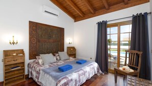 Un dormitorio con techo de madera, una cama grande con colcha estampada, dos mesitas de noche, una butaca y vistas a una piscina a través de un ventanal.
