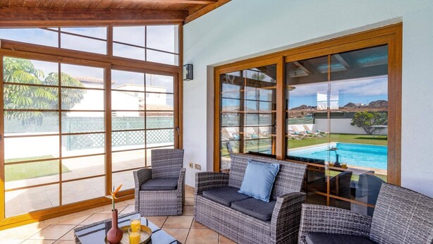 Un patio iluminado por el sol con mobiliario de mimbre, una mesa de vidrio y vista a una piscina.