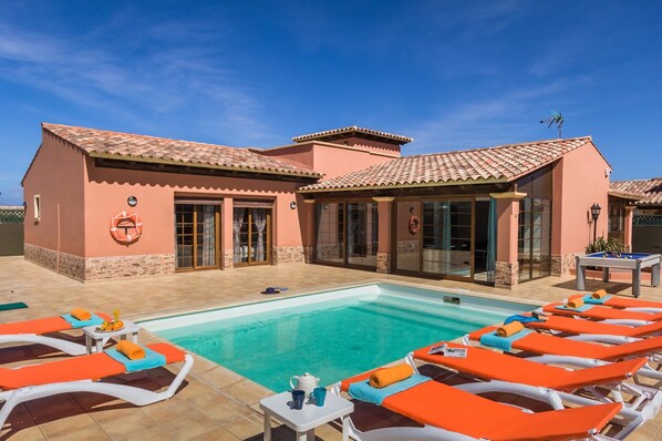 A poolside area with lounge chairs, a pool, and a building with a tiled roof and large windows. - Villa Simona by Villa Plus (Corralejo)