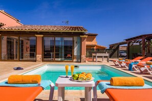 A poolside area with lounge chairs, a table with fruit and drinks, and a building with large windows and a tiled roof. - Villa Simona by Villa Plus (Corralejo)