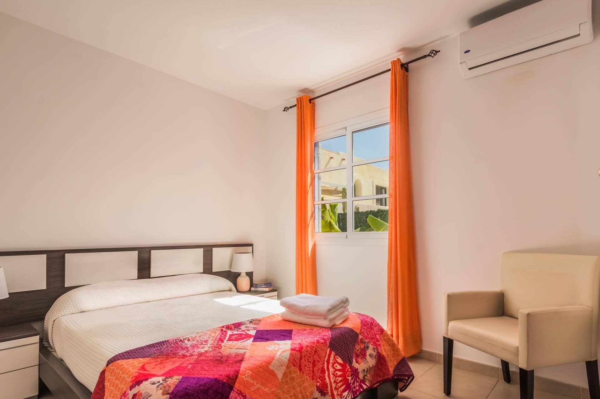 A bedroom with a bed, a chair, an air conditioning unit, and a window with curtains.