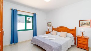 Un dormitorio con cama, mesita de noche, cómoda, ventana con cortinas y dos cuadros en la pared.