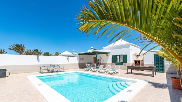 Una piscina con sillas de descanso, zona de barbacoa y un edificio blanco con contraventanas verdes.