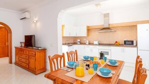 Una cocina moderna con armarios blancos, una mesa de comedor de madera dispuesta con cuencos azules y jugo de naranja, y un televisor sobre un mueble de madera.