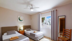 Ein Hotelzimmer mit zwei Betten, einem Deckenventilator, einem Fenster mit Vorhängen, einem Sessel und einem Bild an der Wand.